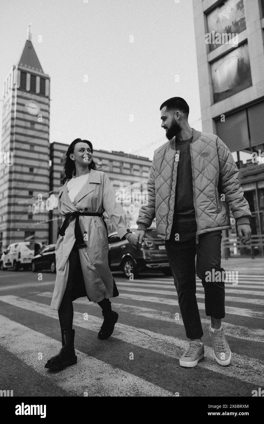 Happy young couple holding each other hands and crossing pedestrian crossing on city street against background of buildings and cars. Black and white Stock Photo