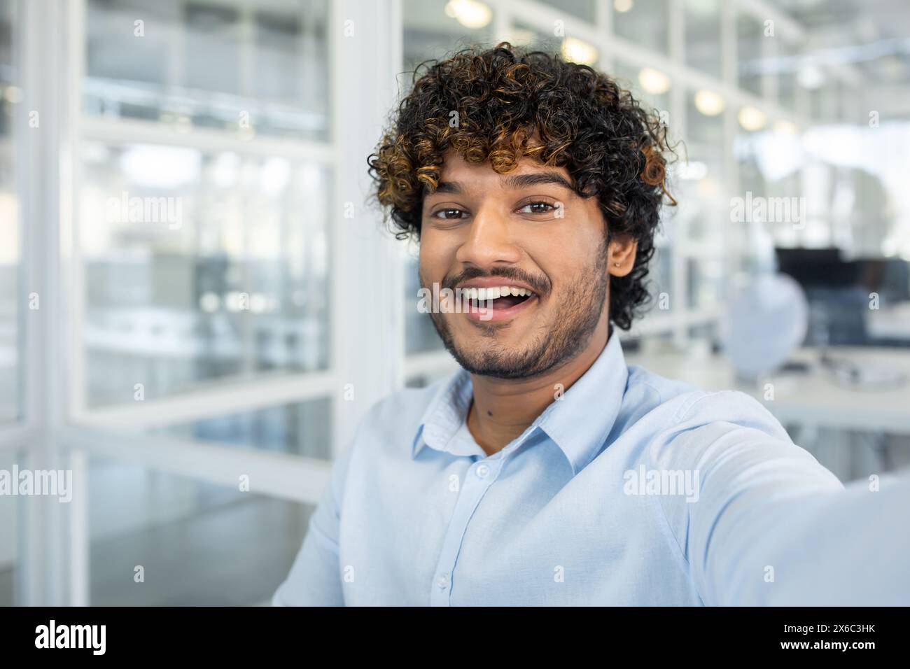 Cheerful young man snapping a selfie in a bright, modern office space, showcasing joy and a professional atmosphere. Stock Photo