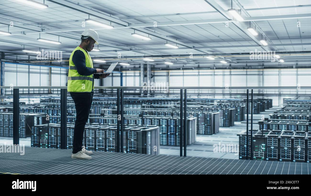 Data Center Engineer in High Visibility Vest Using Laptop Computer. Server Farm Cloud Computing Facility with Black Male System Administrator Working with Data Protection Network for Cyber Security. Stock Photo