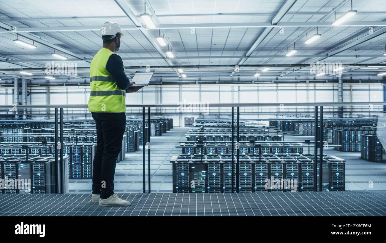 Data Center Engineer in High Visibility Vest Using Laptop Computer. Server Farm Cloud Computing Facility with Black Male System Administrator Working with Data Protection Network for Cyber Security. Stock Photo