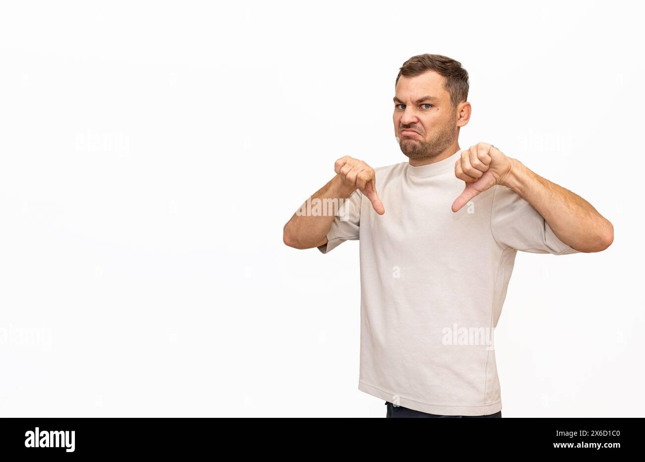Dissatisfied man shows thumbs down on white background in studio. Male person expressing disapproval, disagreement, or dissatisfaction. Stock Photo