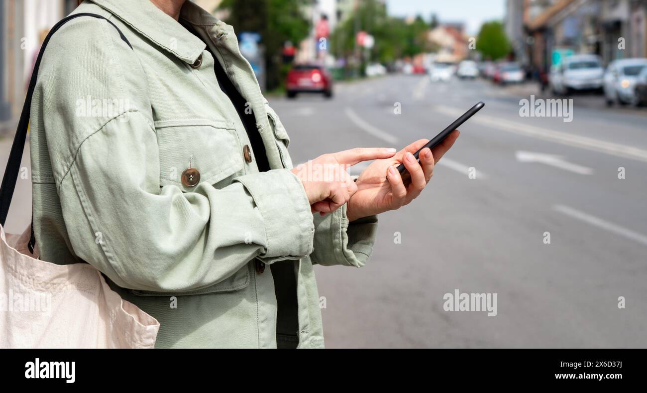 Female urban person stands near the city road and booking a taxi using her mobile app. Free floating car-sharing service online searching. Stock Photo