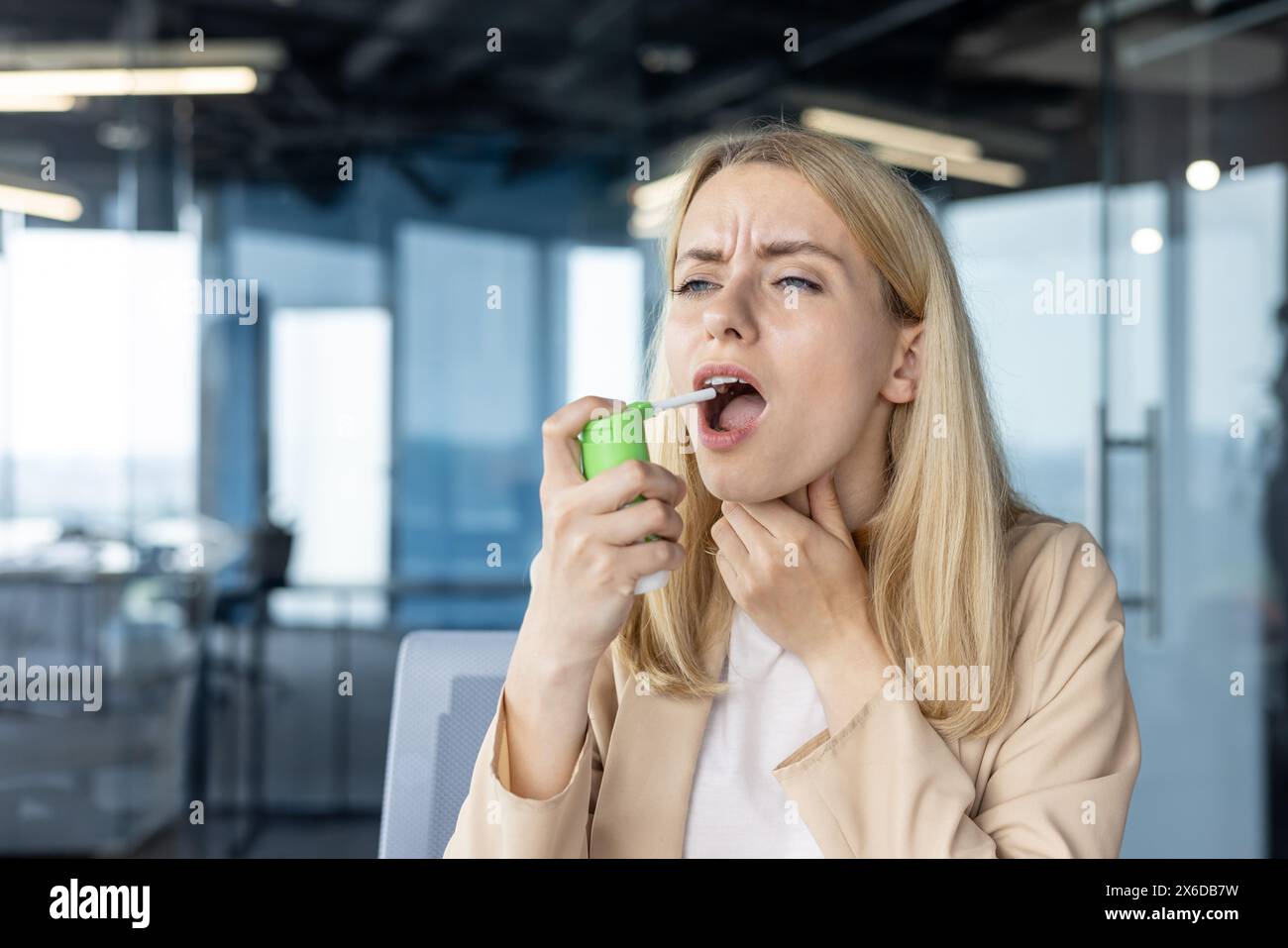 A woman in an office uses a throat spray for sore throat relief. She appears to be in discomfort while medicating in a modern workspace. Stock Photo