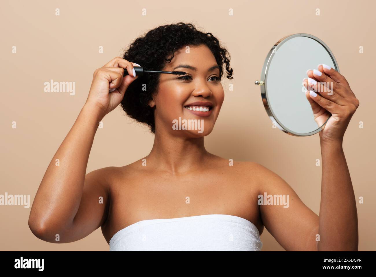 Young Woman Applying Mascara Holding Hand Mirror Against Beige Background Stock Photo