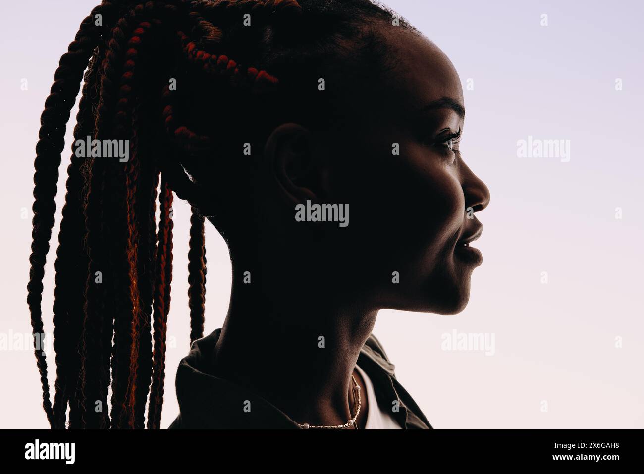 Silhouette of a young woman standing in a studio with braids styled in a ponytail. Her headshot features a closeup sideview of her face looking away, Stock Photo