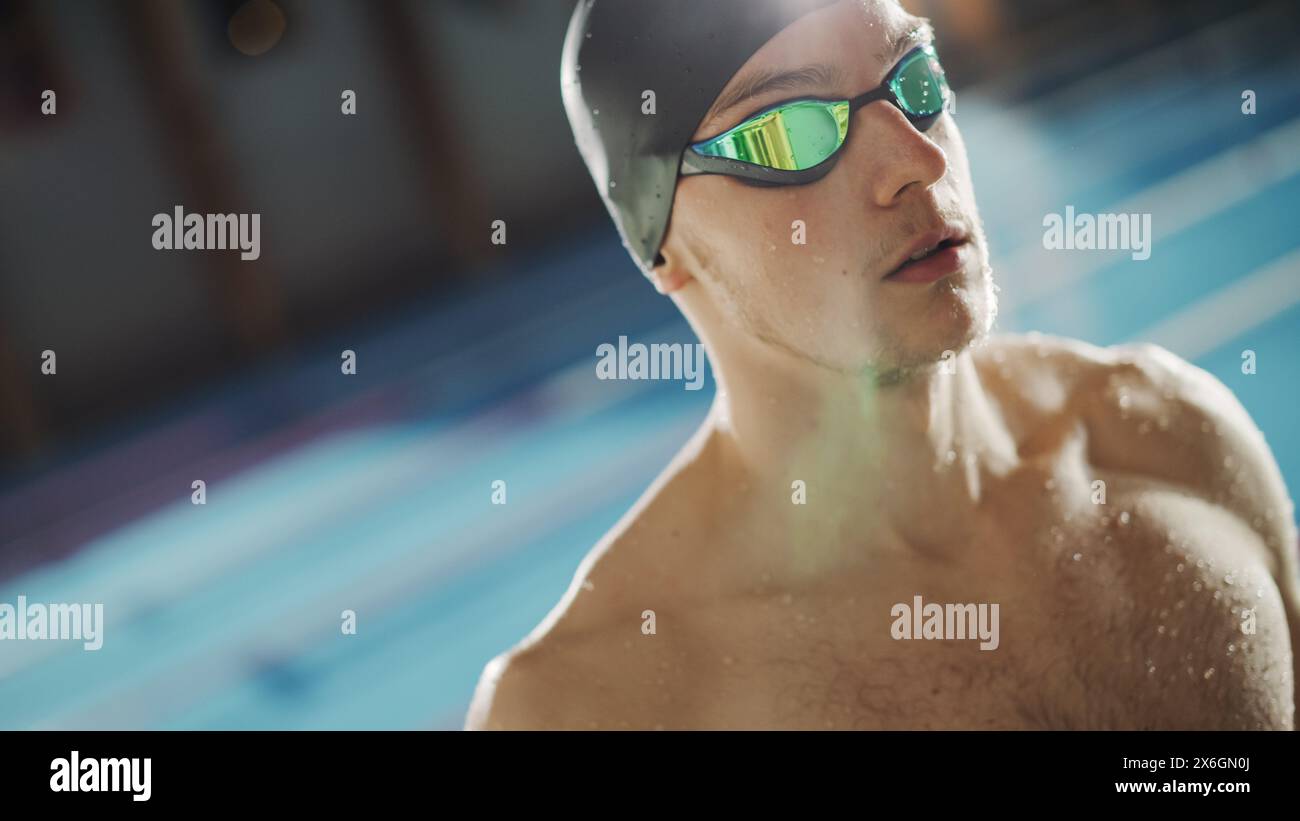 Handsome Professional Male Swimmer in Swimming Pool, Wearing Cap and Glasses, Looks Confidently at the Camera, Ready to Win the Championship. Portrait of Confident Man. Low Dutch Angle Stock Photo