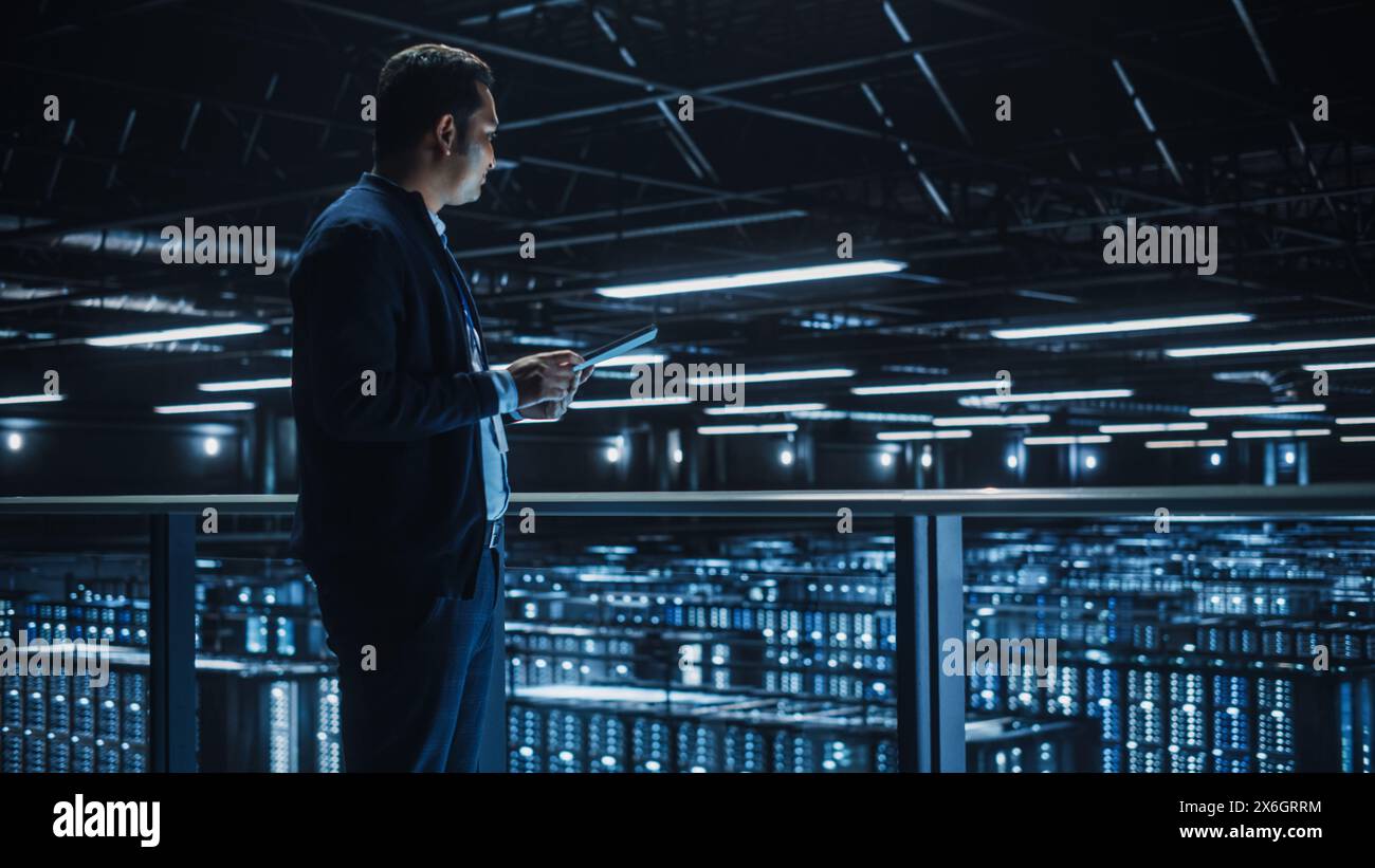 Handsome Smiling IT Specialist Using Tablet Computer in Data Center. Succesful Businessman and e-Business Entrepreneur Overlooking Server Farm Cloud Computing Facility. Stock Photo