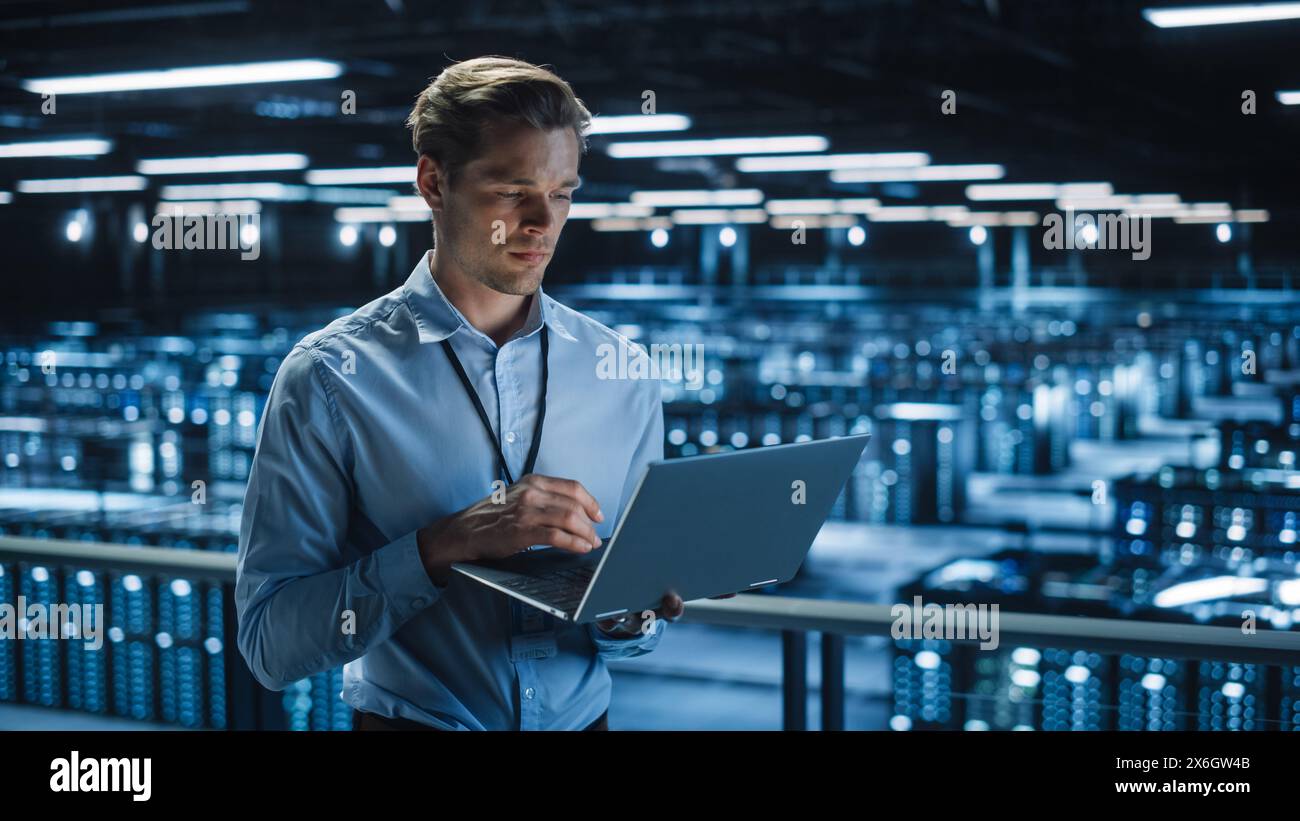 Handsome Smiling IT Specialist Using Laptop Computer in Data Center. Succesful Businessman and e-Business Entrepreneur Overlooking Server Farm Cloud Computing Facility. Stock Photo