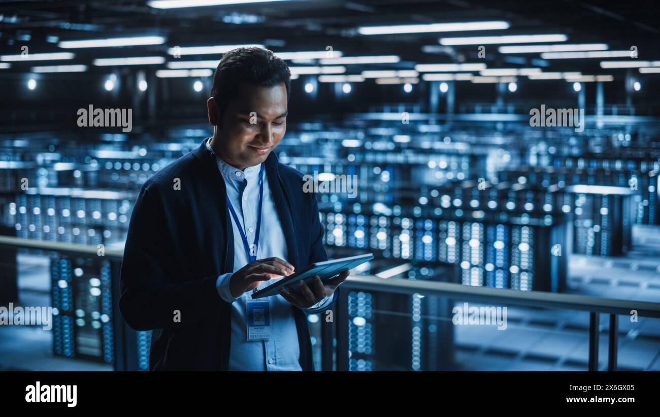 Handsome Smiling IT Specialist Using Tablet Computer in Data Center. Succesful Businessman and e-Business Entrepreneur Overlooking Server Farm Cloud Computing Facility. Stock Photo