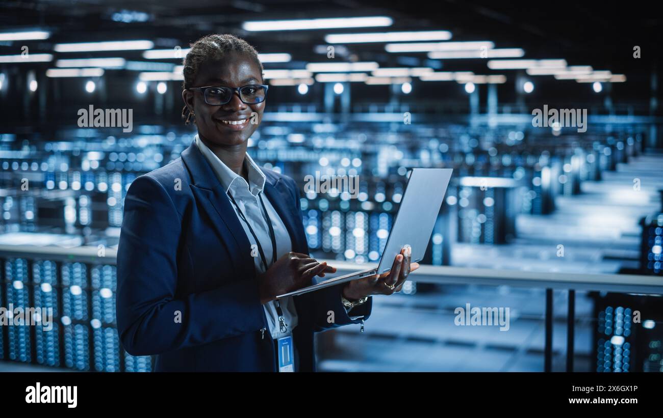 Data Center: Female Programmer Using Laptop Computer, Smiling and Looking at Camera. In Cloud Computing Server Farm System Administrator Working on IT Maintenance for Iaas, saas, paas. Stock Photo