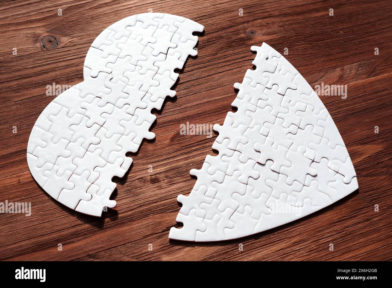 White heart-shaped puzzle, split in half, lying on a textured wooden table. Separation, longing or incomplete love related concept. Stock Photo