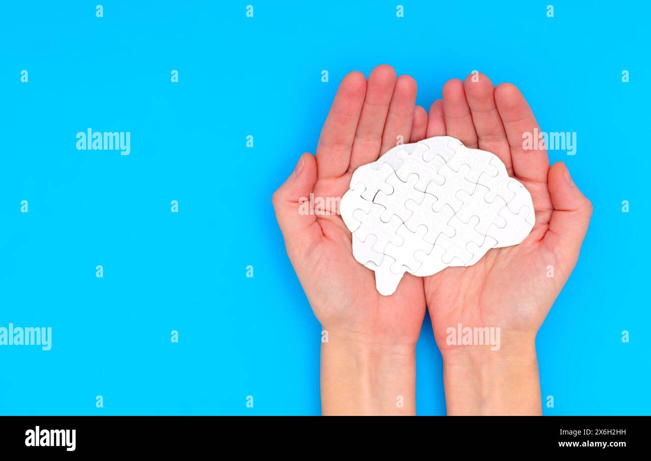 Hands delicately hold a white puzzle piece intricately shaped like a brain against a serene blue backdrop. Cognition, intelligence and mental acuity c Stock Photo
