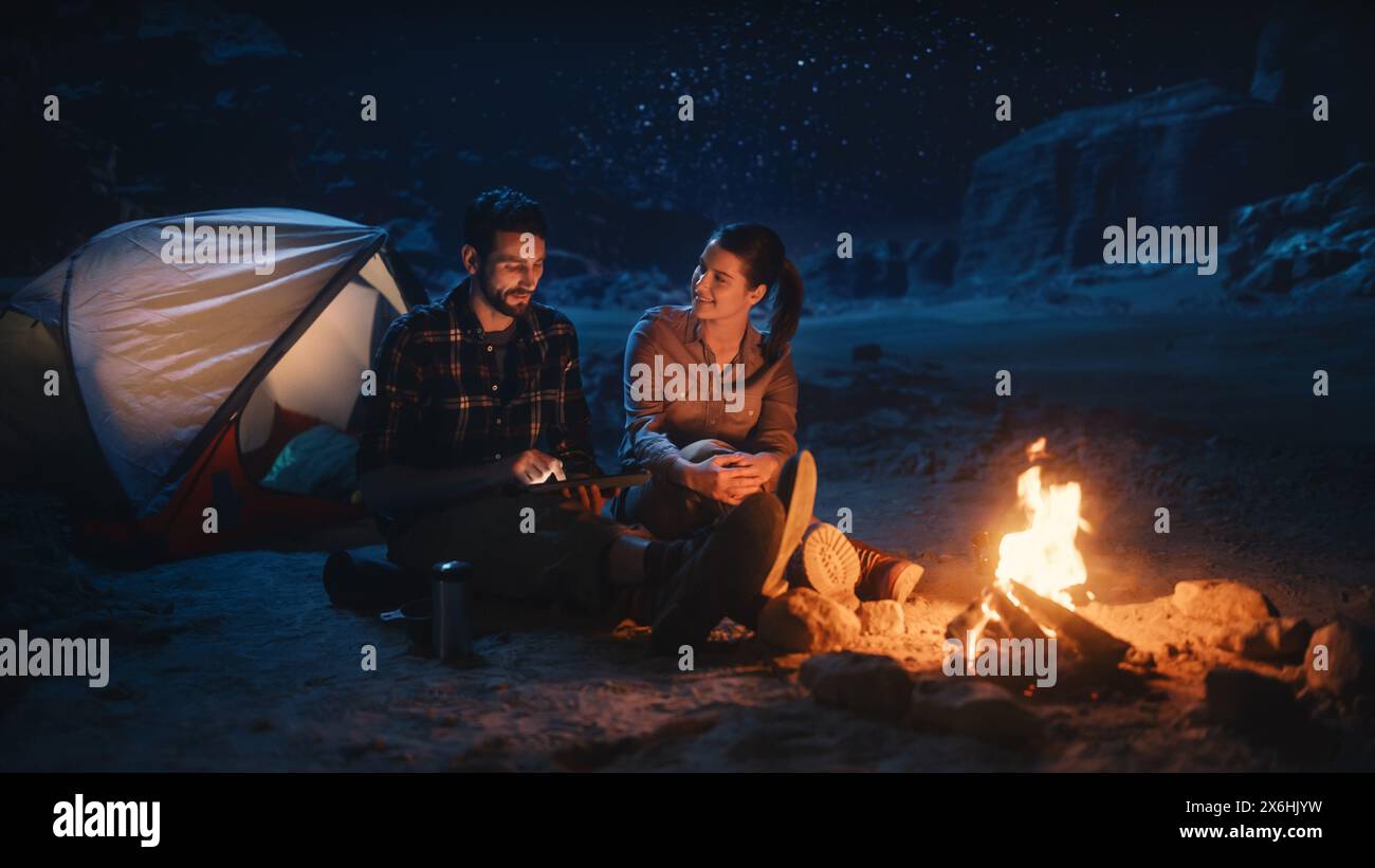 Happy Couple Nature Camping in the Canyon at Night, Use Digital Tablet Computer, Sitting by Campfire. Two people Have Fun, Smile, Post Traveling Photos on Social Media, Watch Funny Videos on Internet Stock Photo