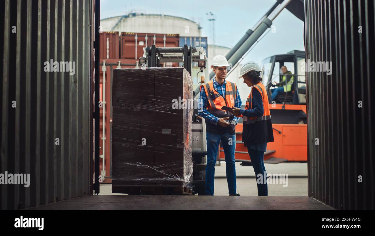 Forklift Driver Loading Shipping Cargo Container with Full Pallet with Boxes in Logistics Port Terminal. Latin Female Industrial Supervisor and Safety Inspector with Tablet Talking, Managing Process. Stock Photo