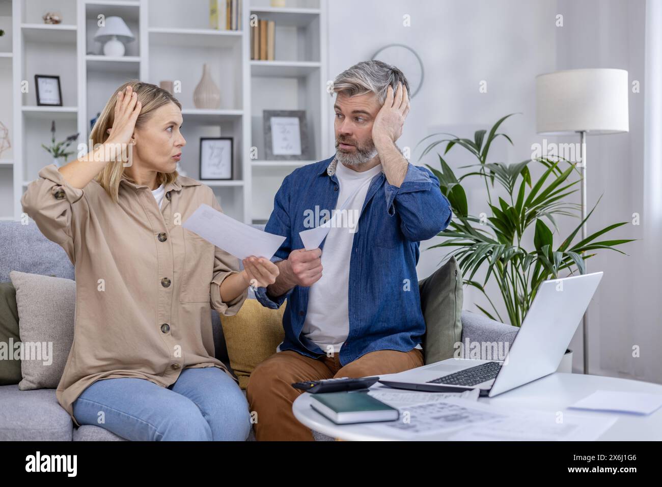 Stressed couple sitting on the couch discussing bills and finances. They appear worried while holding papers in their hands and looking at each other. Stock Photo