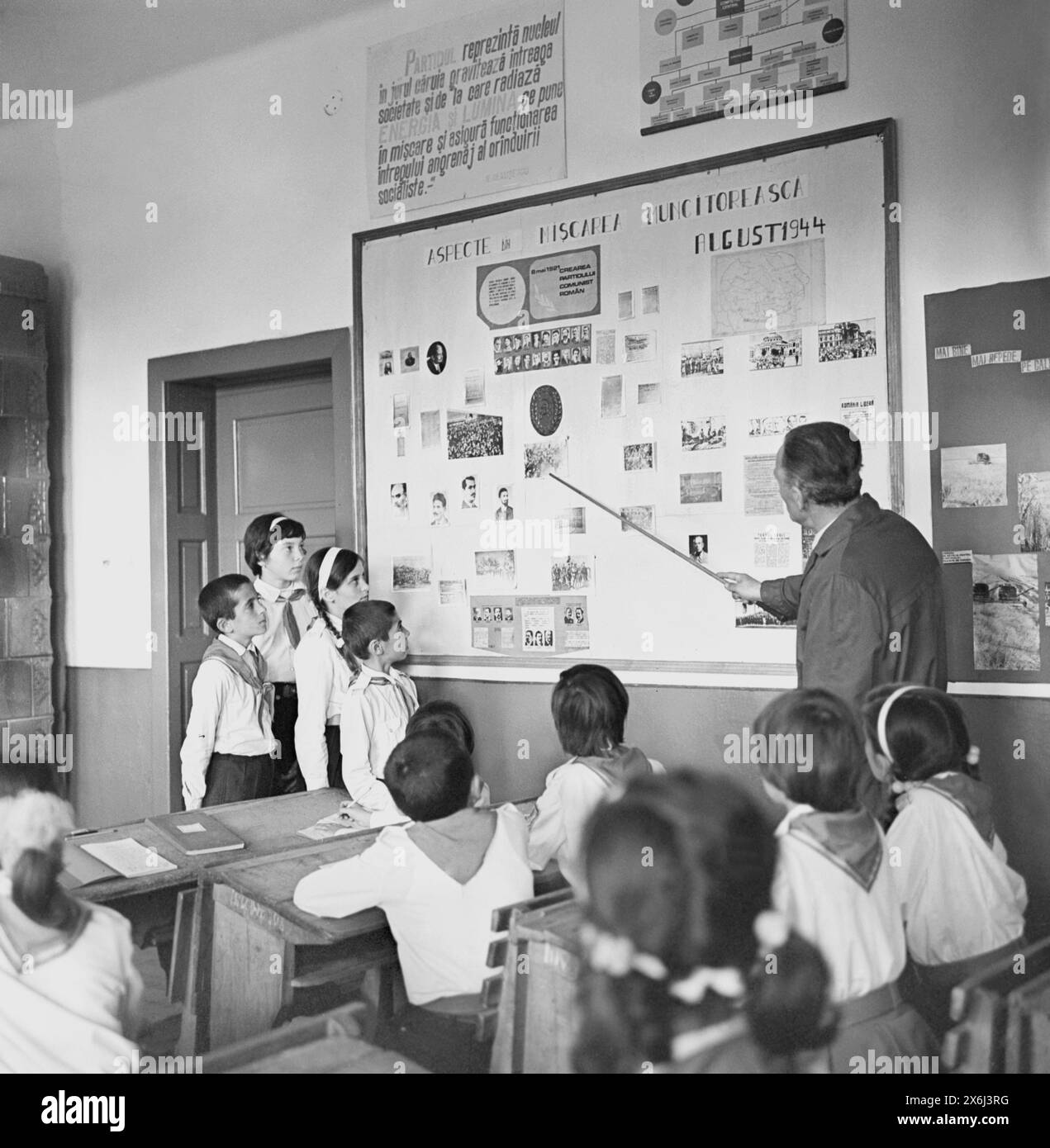 Socialist Republic of Romania, 1970s. Elementary students during a class in a governmental school. The teacher is pointing to a propagandist board entitled 'Aspects of the Worker's Movement', including the creation and the symbols of the Communist Party, its main leaders, & the events of August 1944, which led to Romania getting under the Soviet control. Another board at the top says: 'The Party represents the nucleus around which the whole society gravitates and from which radiates the energy and light that sets in motion and ensures the functioning of the entire gear of the Socialist system. Stock Photo