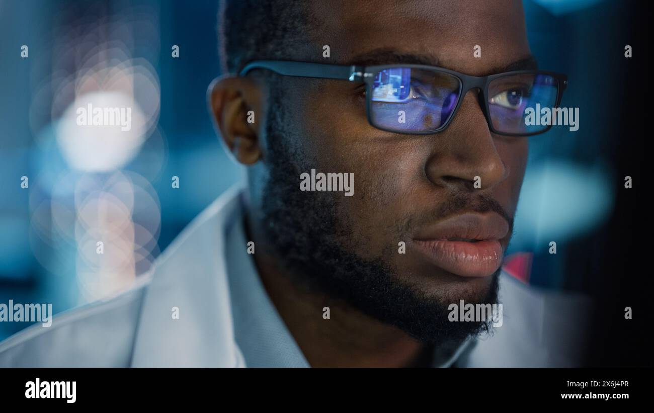 Portrait of Handsome Black Man Wearing Glasses Working Confidently on a Computer. Young Intelligent Male Engineer or Scientist Working in Laboratory. Bokeh Blue Background. Close-up Shot Stock Photo