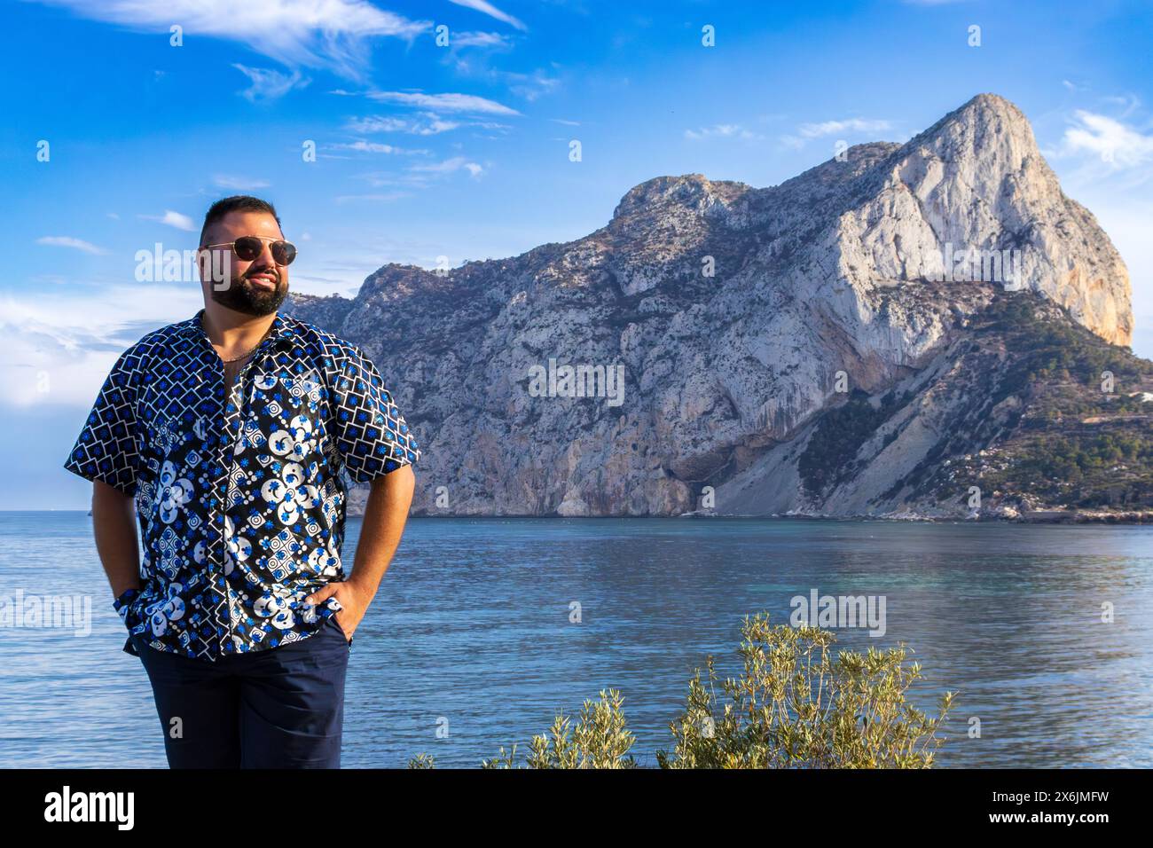 This portrait captures a 30-year-old man as he stands in serene contemplation, with the majestic Ifach Mountain of Calpe, Spain. Stock Photo