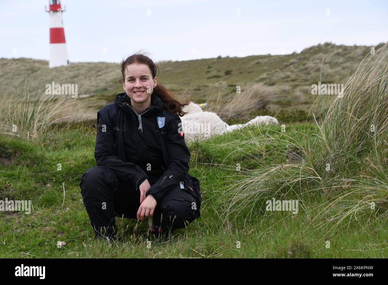 List, Germany. 09th May, 2024. Ranger Stella Kinne kneels in front of two sheep and the east lighthouse in the Lister Ellenbogen nature reserve (Sylt). The 26-year-old is responsible for protecting the animals here in the nature reserve. Credit: Lea Sarah Albert//dpa/Alamy Live News Stock Photo