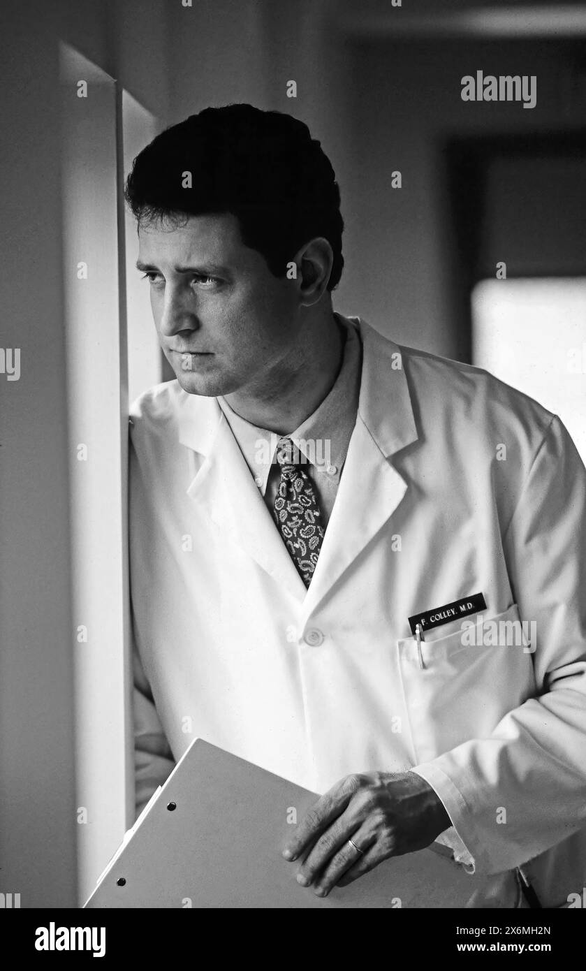 A doctor in a white coat stands thoughtfully by a window in a hospital corridor. He appears reflective, holding a folder and looking out towards the l Stock Photo