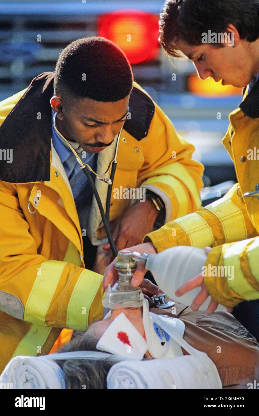 Two dedicated paramedics in yellow jackets provide urgent medical assistance to an injured individual on the street. Using a stethoscope and other ess Stock Photo