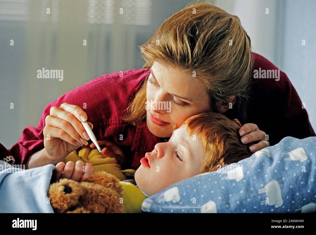 A mother tenderly checks her childs temperature with a thermometer as they rest in bed. The child clutches a teddy bear and appears unwell while recei Stock Photo