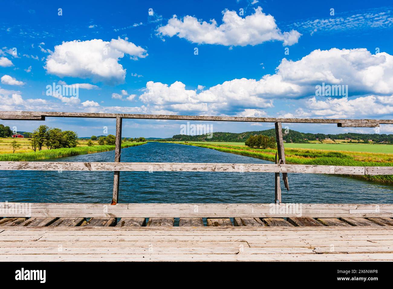 A rustic wooden bridge spans a calm river under a bright blue sky scattered with fluffy white clouds. Verdant fields and trees surround the water, cre Stock Photo