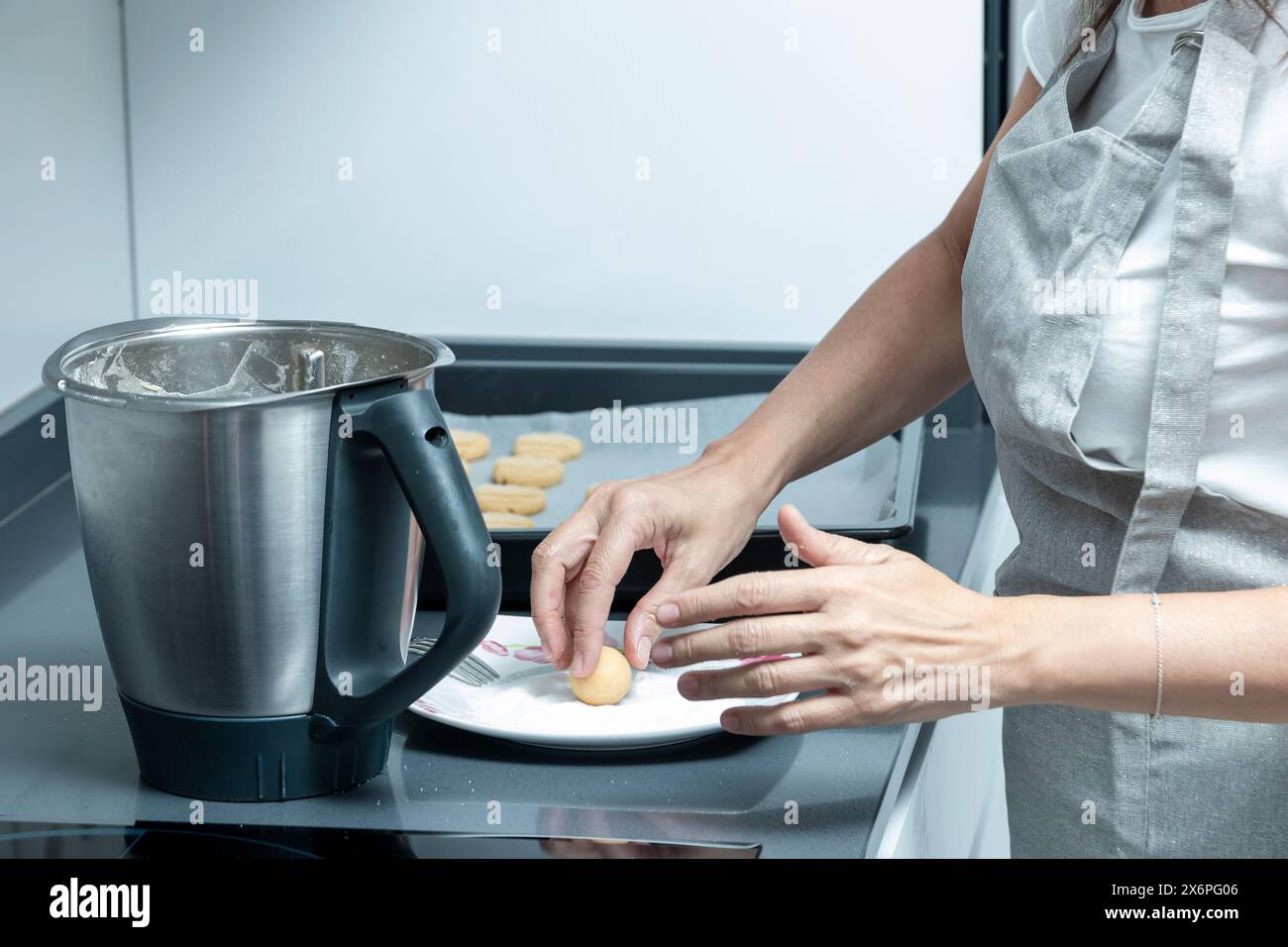 person in an apron placing shaped cookie dough onto a baking sheet next to a mixing bowl in a modern kitchen Stock Photo