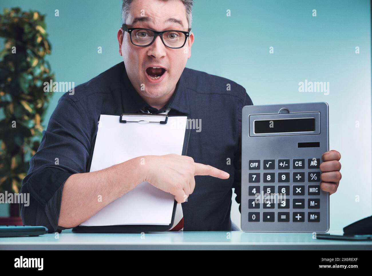 Man in dark shirt and glasses, holding a clipboard with one hand and pointing at a large calculator with the other, expresses surprise, seated at a de Stock Photo