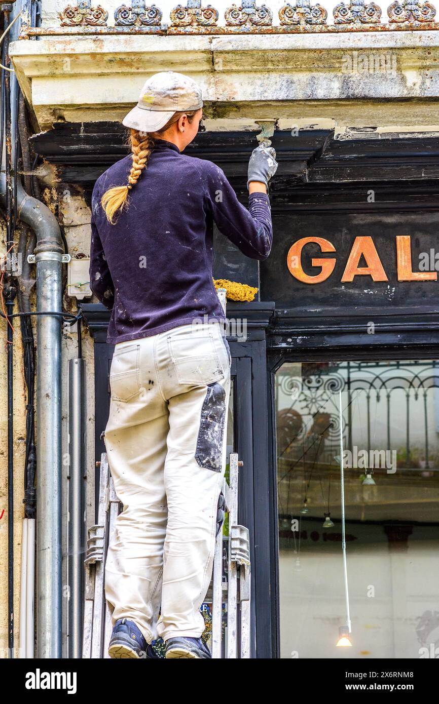 Young female decorator sanding shop front prior to repainting - Loches, Indre-et-Loire (37), France. Stock Photo