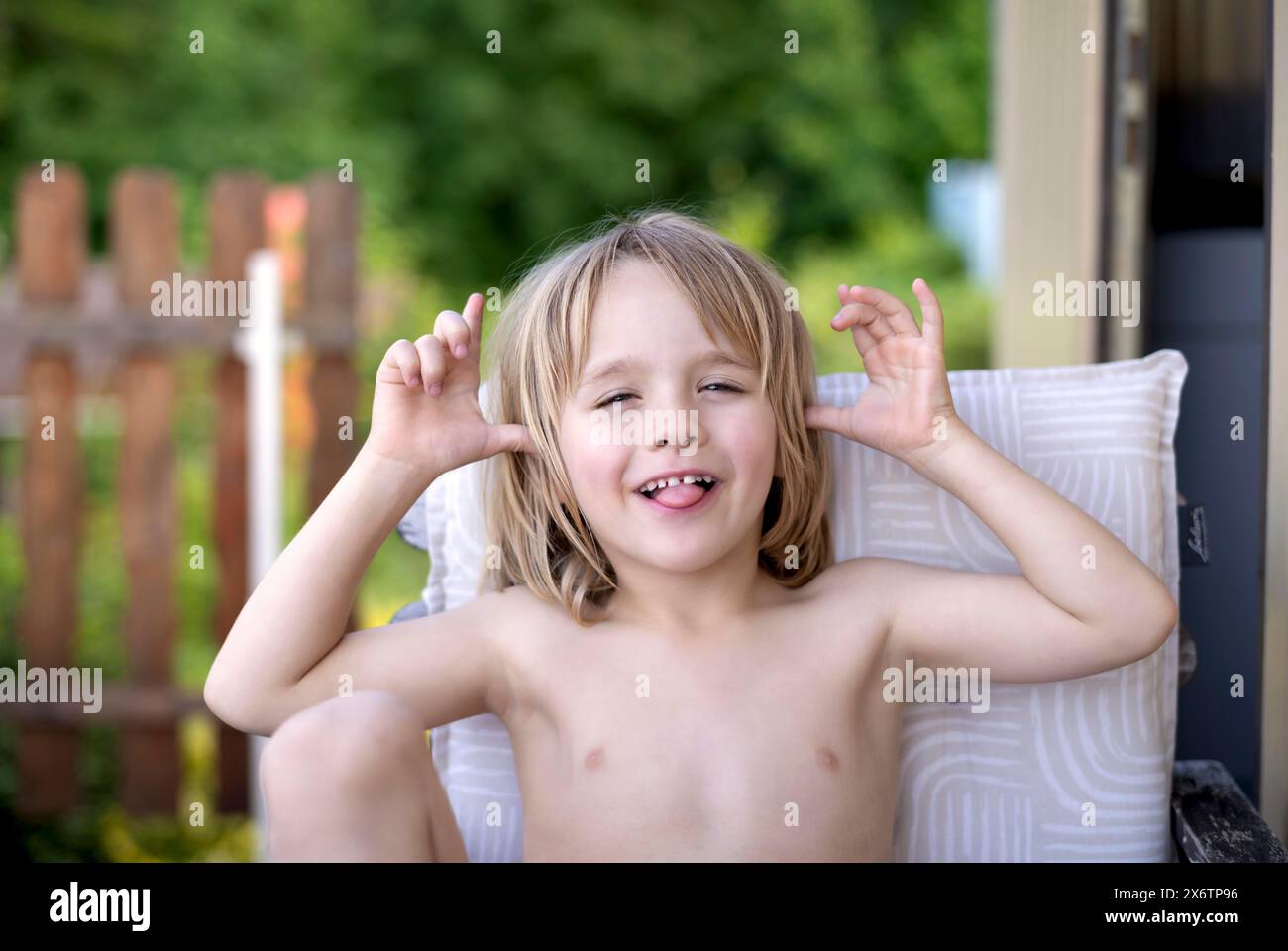 Boy, 5 years, blonde, sitting in garden chair, garden, grimacing, sticking out tongue, summer, Stuttgart, Baden-Wuerttemberg, Germany Stock Photo