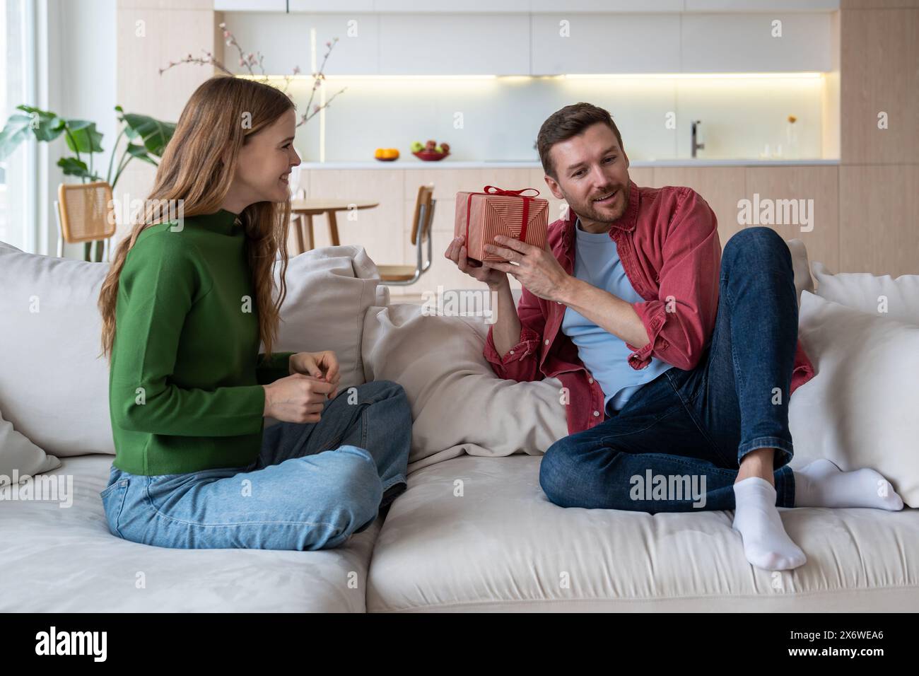 Romantic merry couple sitting on sofa, celebrating family event, holding giftbox, guessing content Stock Photo