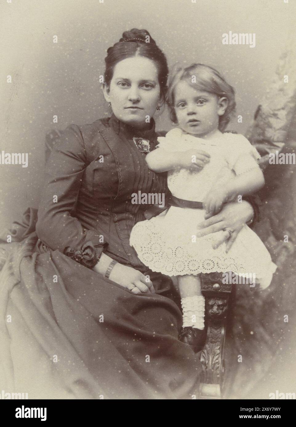 Studio portrait of mother and daughter, Studio portrait of mother and daughter. The mother wears a black high-necked dress with a collar closed with a brooch. The daughter wears a white dress and they sit together on a chair., photograph, Kameke, (mentioned on object), The Hague, 1890 - 1920, paper, height, 135 mm × width, 95 mm, height, 260 mm × width, 210 mm Stock Photo