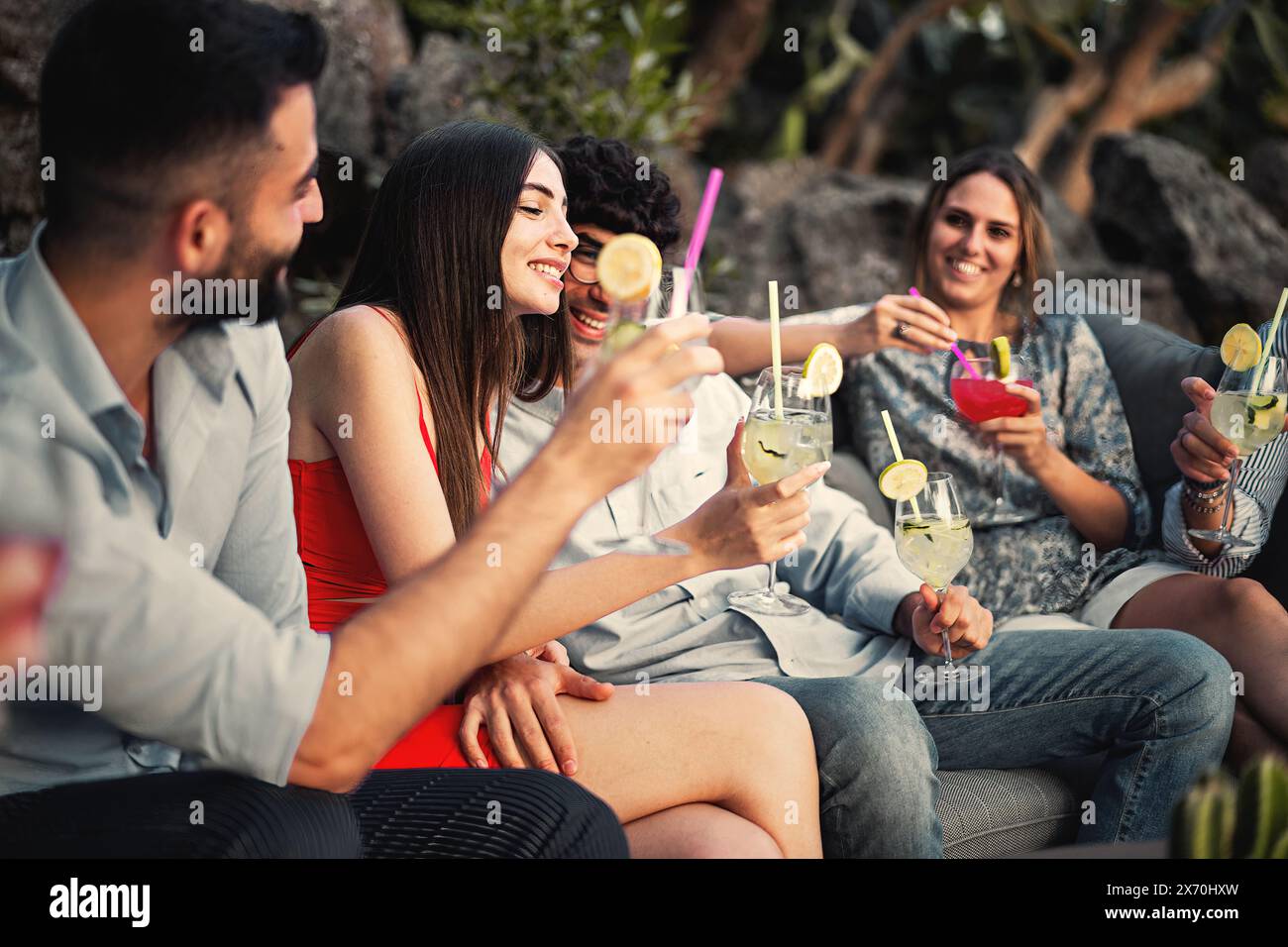 A group of friends enjoying a fun evening outdoors, toasting with refreshing cocktails. They are seated in a comfortable, relaxed setting, sharing lau Stock Photo