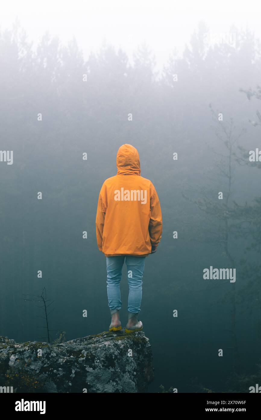 Bulgaria - 05 22 2024: Man with vibrant orange jacket and jeans standing on the edge of a rock looking at the bright light over the trees with a lot o Stock Photo