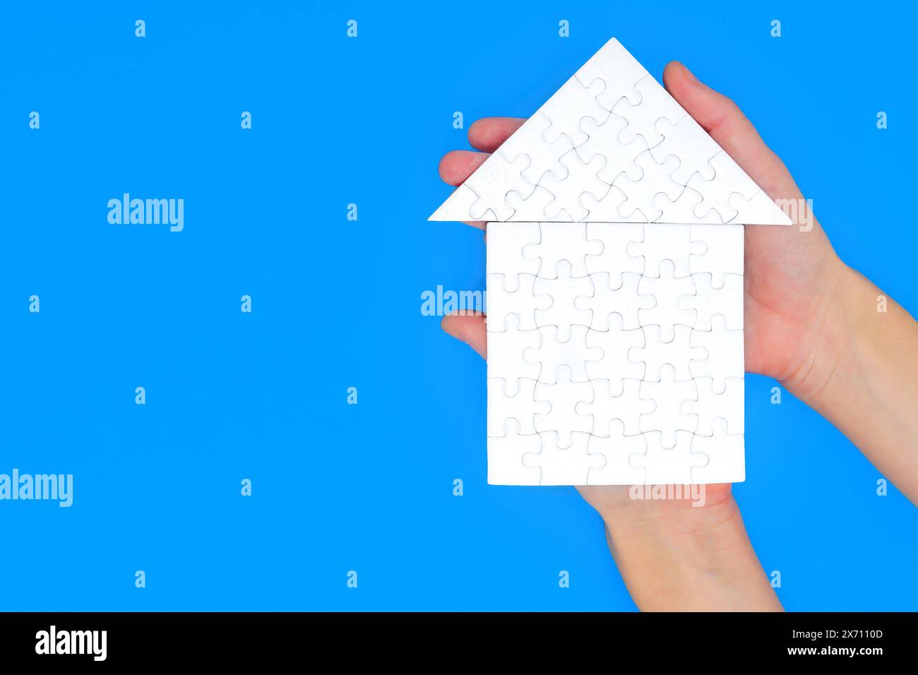 White hip roof house made up of jigsaw puzzle pieces held in both hands against pristine blue background with copy space. Stock Photo