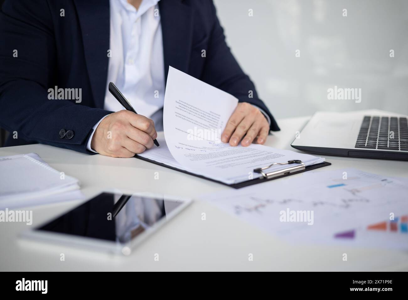 Asian mature man in a business suit signing important documents at an office desk with a laptop, tablet, and papers. Stock Photo