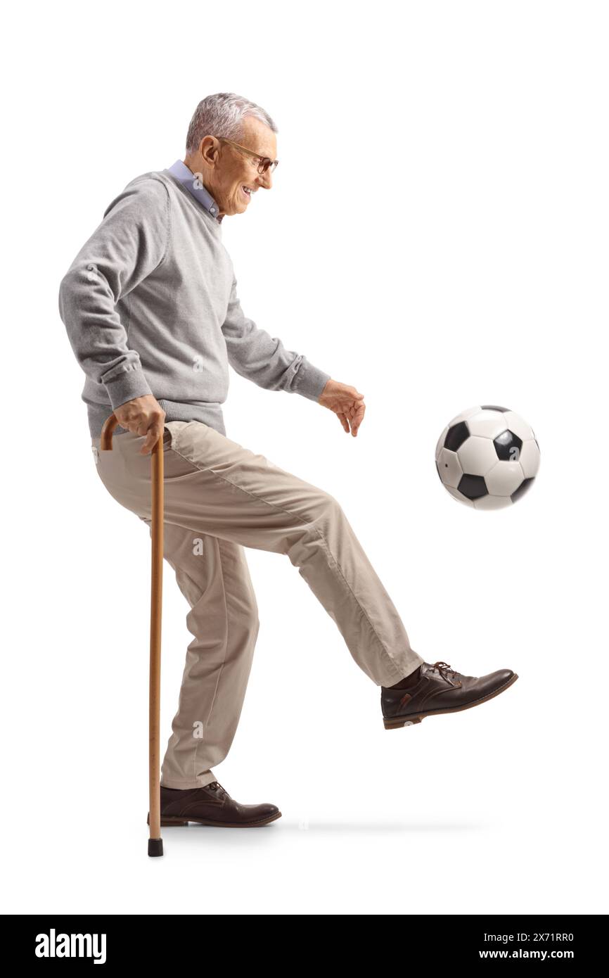 Elderly man leaning on a walking cane and kicking a football isolated on white background Stock Photo