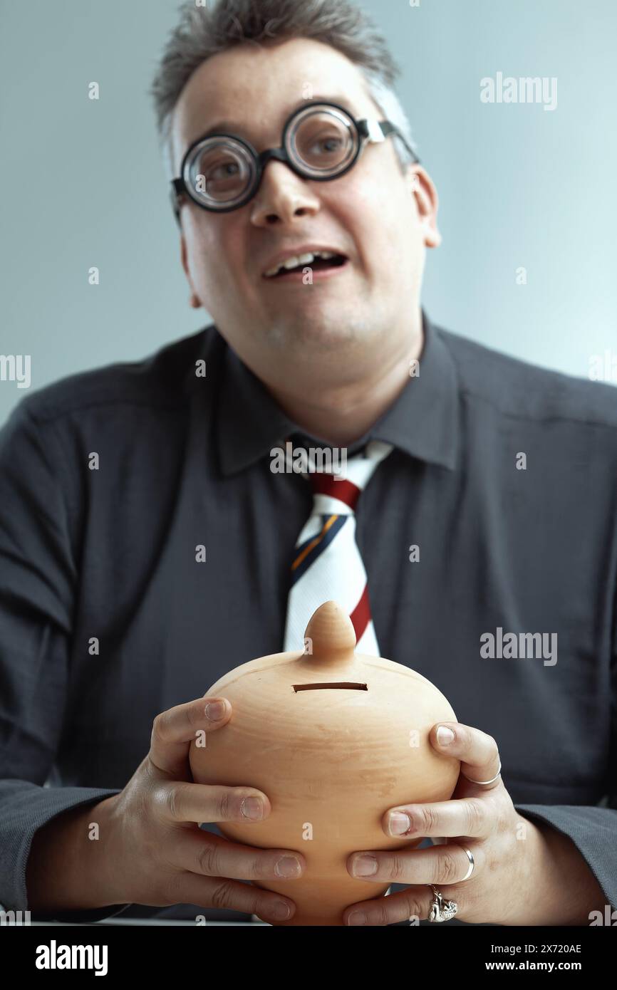 Middle-aged man in dark gray shirt and striped tie, with spiky gray hair and oversized round glasses, awkwardly smiles while holding a ceramic piggy b Stock Photo