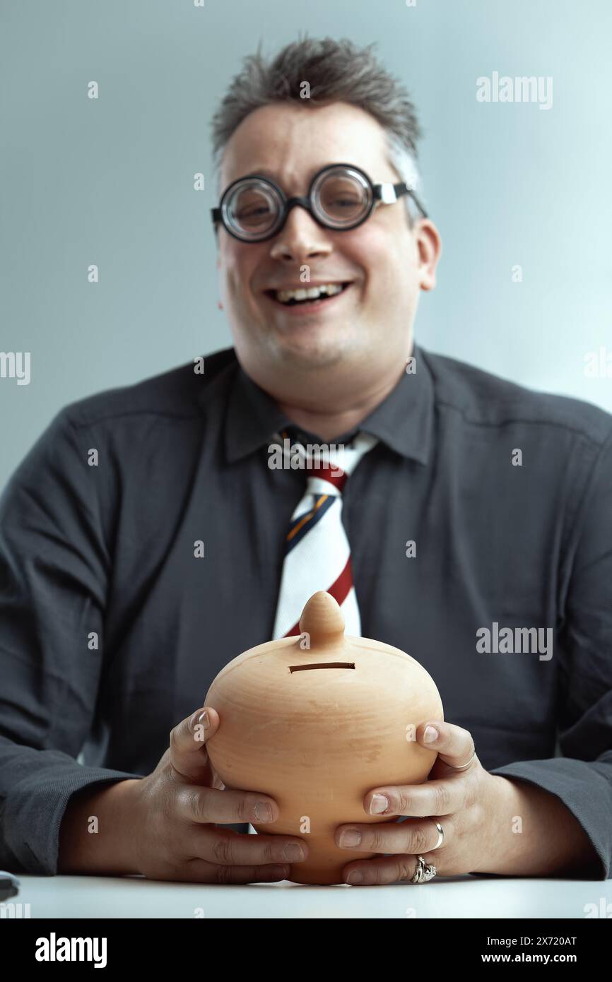 Man with spiky gray hair, dark gray shirt, and striped tie, wearing oversized round glasses, smiles awkwardly while holding a ceramic piggy bank, repr Stock Photo