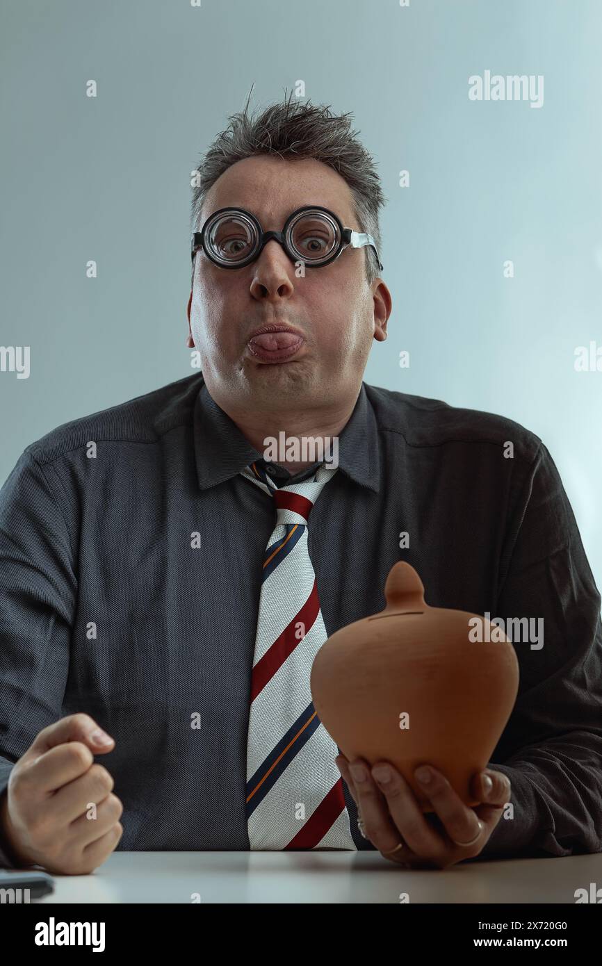 Awkward man in a dark gray shirt and striped tie, with spiky gray hair and oversized round glasses, holds a piggy bank, representing poor money manage Stock Photo