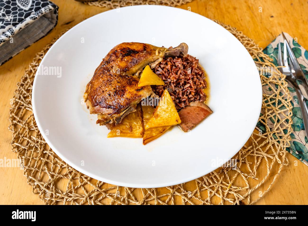 Baked chicken leg with red rice, pineapple piece and bacon in white plate on wooden table, serving size, closeup. Stock Photo