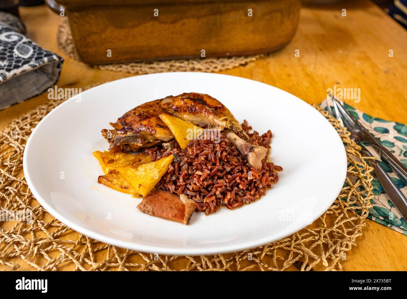 Baked chicken leg with red rice, pineapple pice and bacon on white plate, baking pan,cutlery and potholder on wooden table. Stock Photo