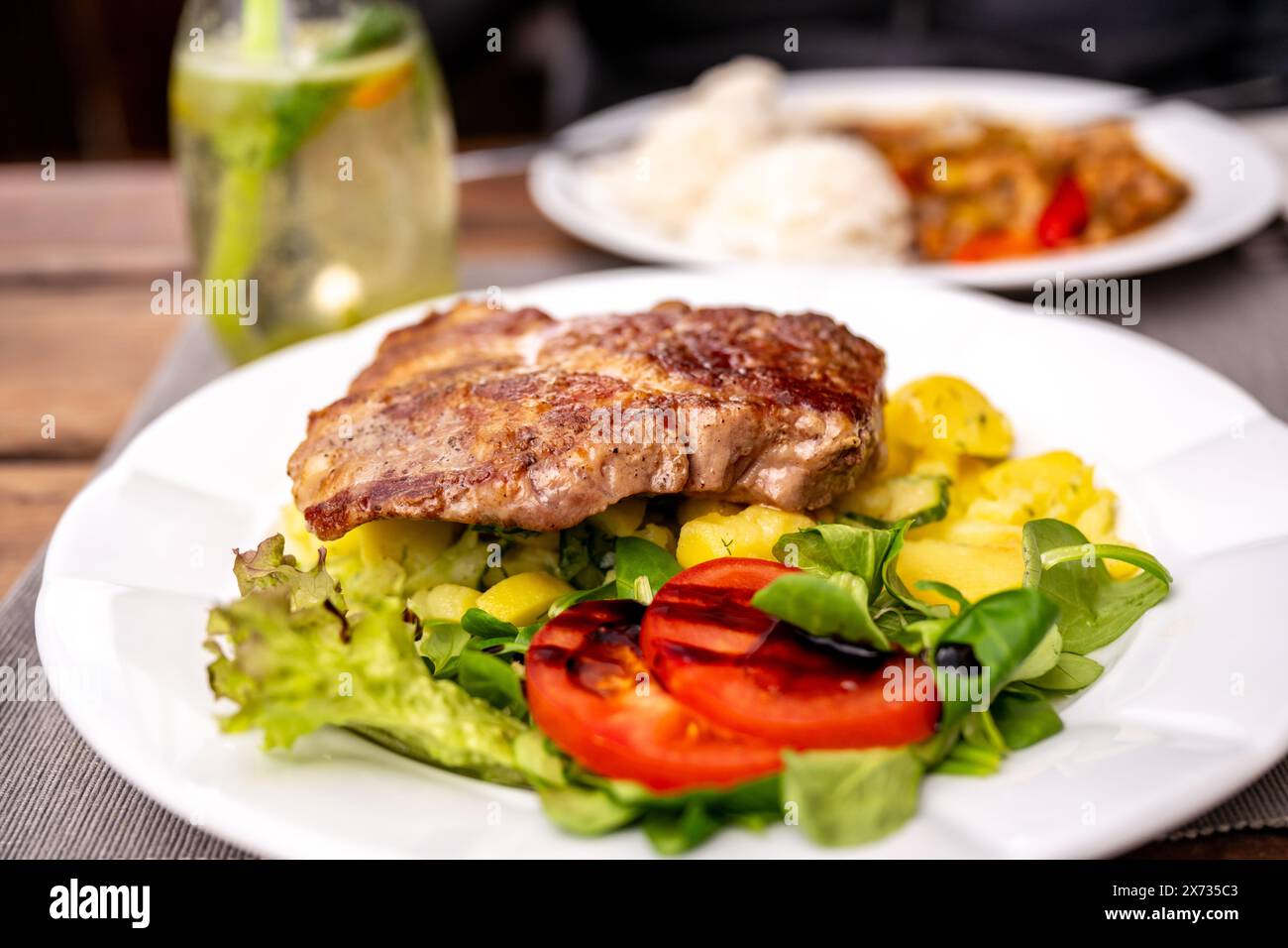 Pork steak on potato salad with vegetable on first white plate, drink in glass and second plate with rice, chicken meat and red pepper on background o Stock Photo