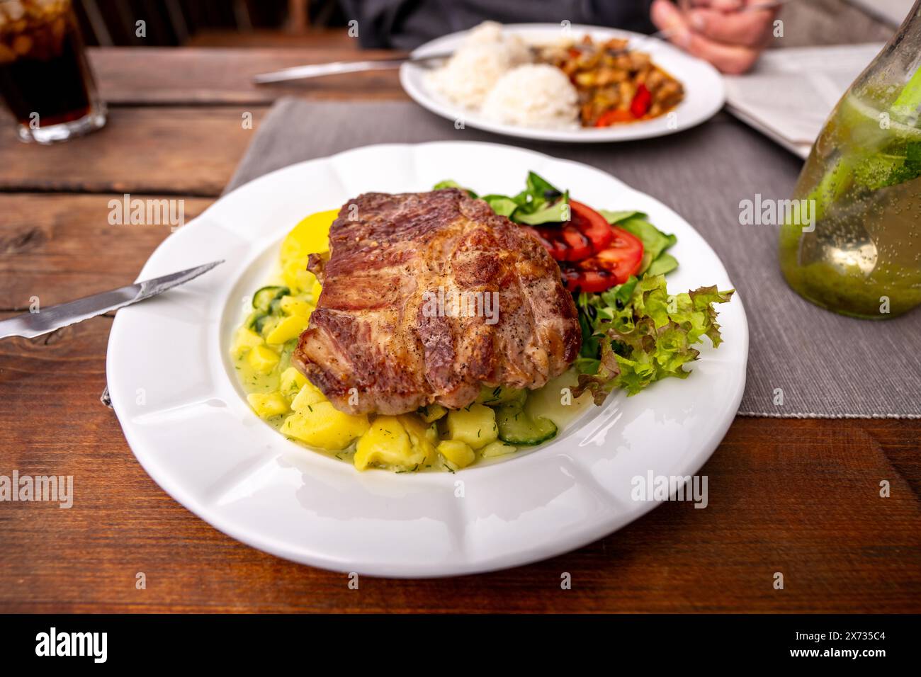 Juicy pork steak with light potato salad and vegetable on white plate, second plate on background with rice, chicken meat with red pepper, drink, knif Stock Photo