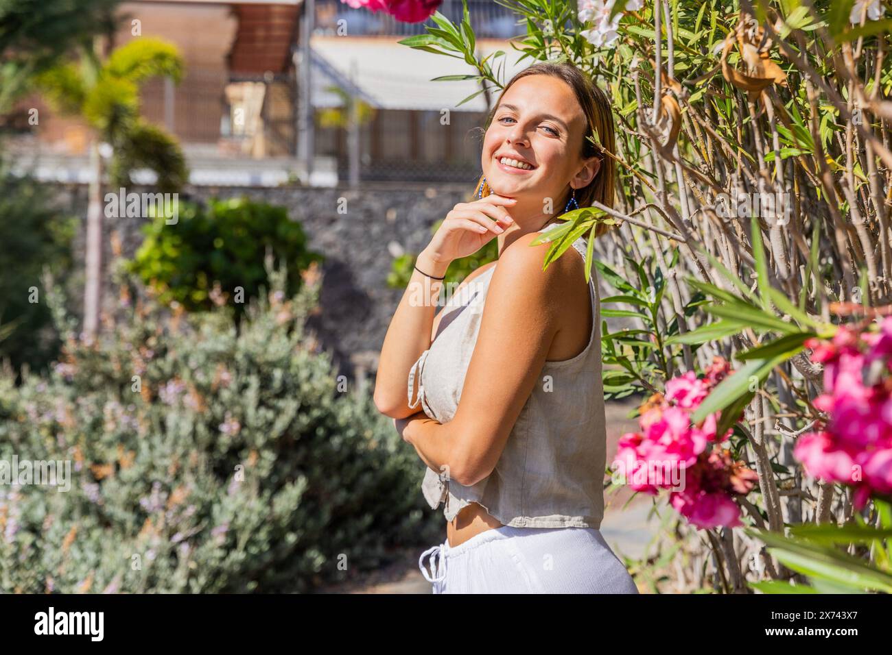 A woman is standing in front of a wall with pink flowers. She is smiling and looking at the camera Stock Photo