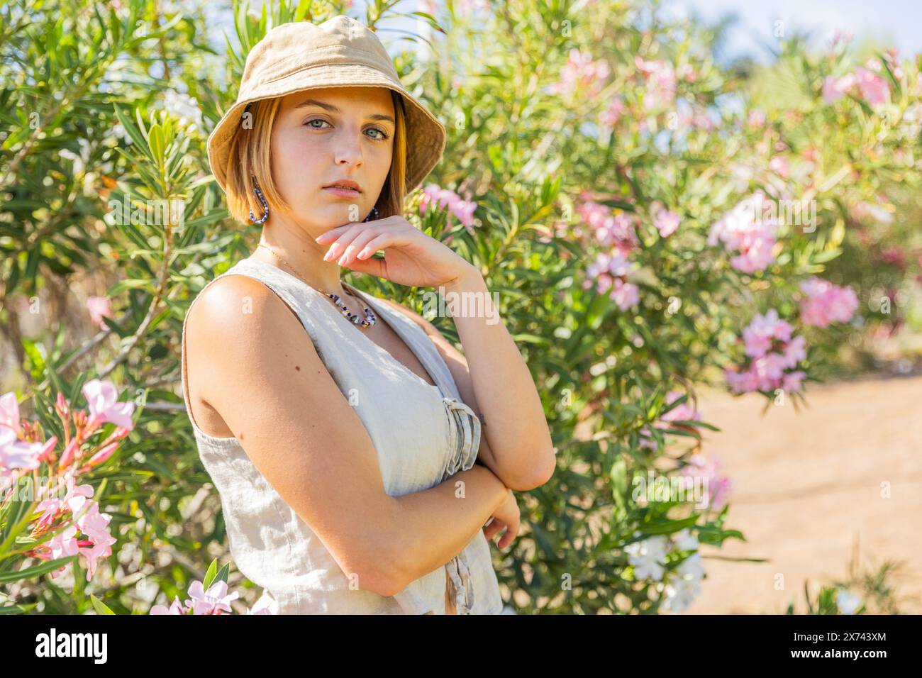 A woman is standing in a field of pink flowers, wearing a hat and a white shirt. She is looking at the camera with a thoughtful expression Stock Photo