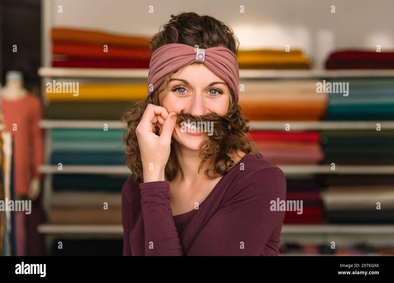 A cheerful fashion designer makes a playful mustache with her curly hair, wearing a stylish mauve headband, against a backdrop of colorful fabric roll Stock Photo