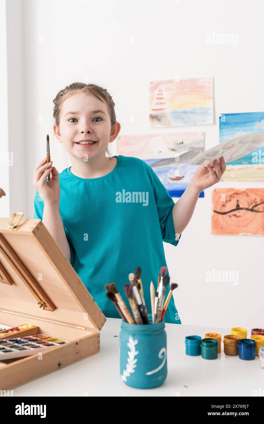 Cute little girl painting with brush and paints with easel and palette ...