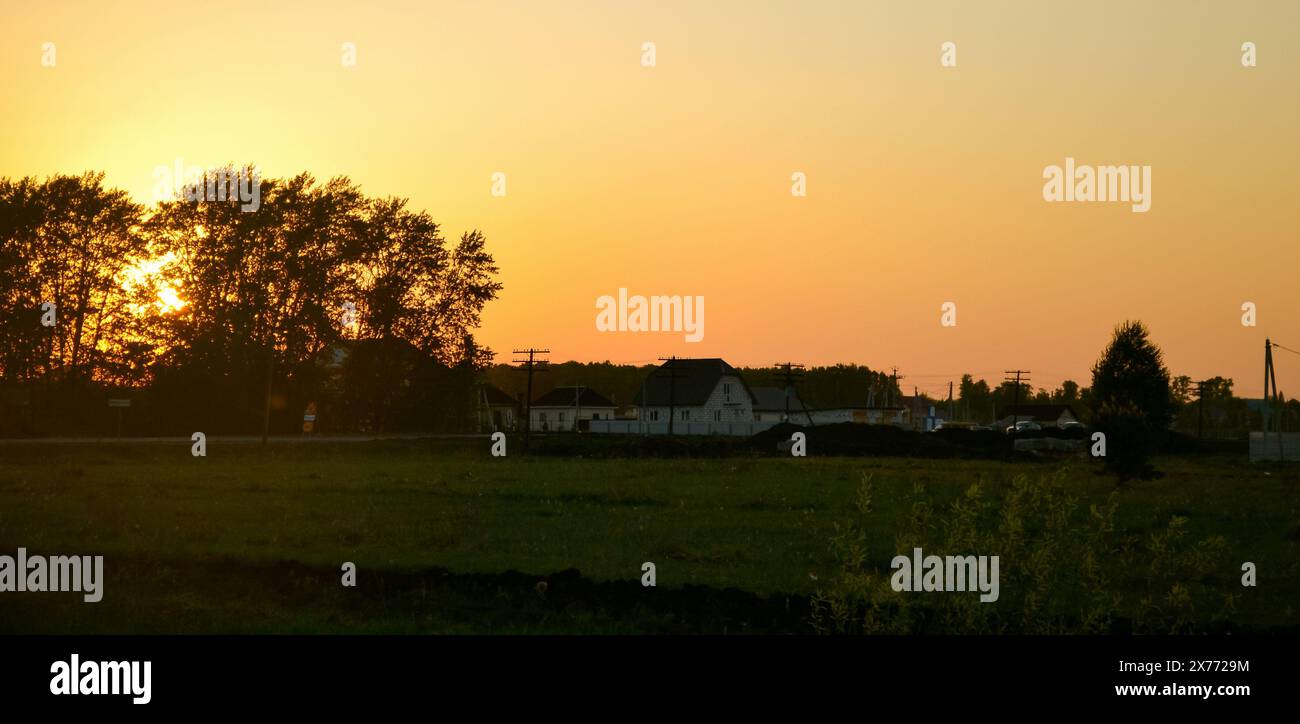 The setting sun behind the silhouettes of trees and the yellow evening sky over the houses in the village Stock Photo